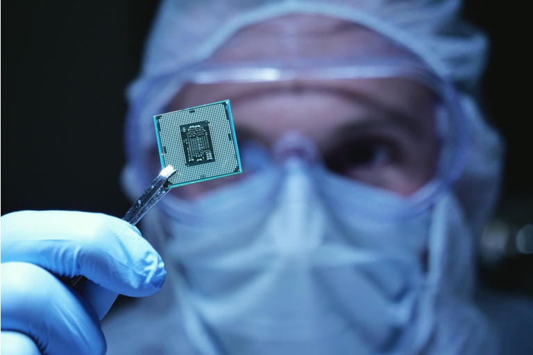 in Ultra Modern Electronic Manufacturing Factory Design Engineer in Sterile Coverall hält Mikrochip mit Symbolen in futuristischer Holographie.